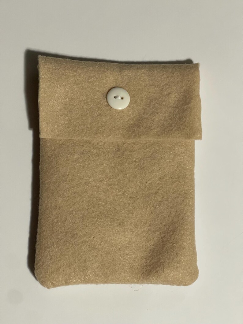 Puede incluir: Una bolsa de fieltro color beige con una solapa cerrada con un bot&oacute;n blanco. La bolsa rectangular parece hecha a mano. El fieltro tiene un aspecto suave y texturizado.