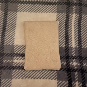 Puede incluir: Una almohada rectangular beige sobre una manta a cuadros. La manta tiene un patr&oacute;n de l&iacute;neas blancas, grises y negras. La almohada est&aacute; hecha de un material suave y texturizado.