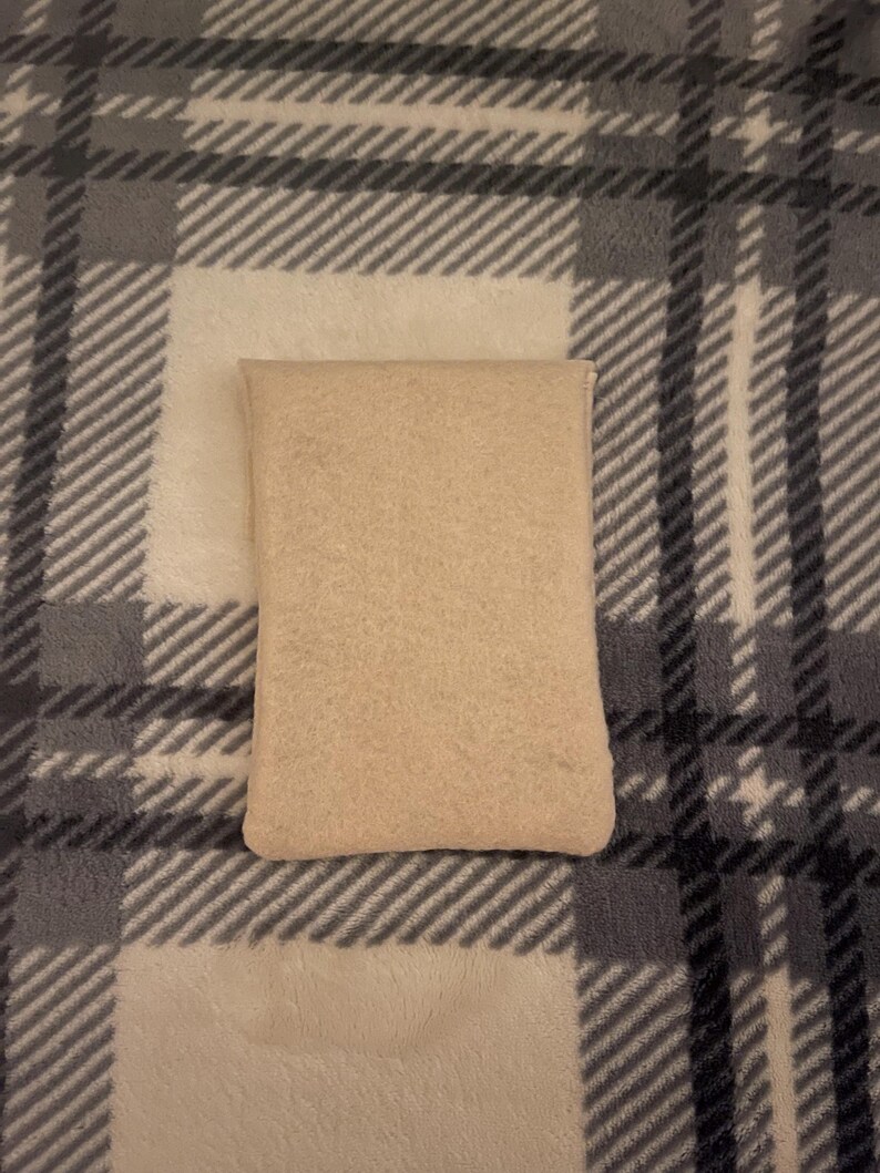 Puede incluir: Una almohada rectangular beige sobre una manta a cuadros. La manta tiene un patr&oacute;n de l&iacute;neas blancas, grises y negras. La almohada est&aacute; hecha de un material suave y texturizado.