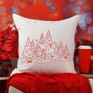 Puede incluir: Almohada blanca con un diseño bordado en rojo de un pueblo de invierno con casas y árboles. La almohada está sobre una manta roja con una linterna blanca y una taza roja en el fondo.
