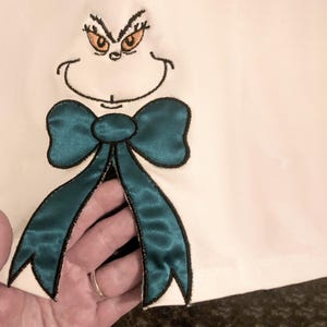 May include: White fabric with a green bow and a cartoon face with orange eyes embroidered on it.