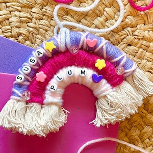 May include: A pink, purple, and white yarn rainbow with the word "PLUM" spelled out in colorful beads. The rainbow has white tassels and is decorated with small hearts and stars.