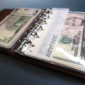 Matte vellum divider tab labeled JANUARY sitting over a clear cash envelope holding US dollar bills in a silver ring binder showing the envelope fit size, on a dark gray surface
