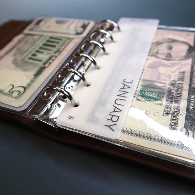 Matte vellum divider tab labeled JANUARY sitting over a clear cash envelope holding US dollar bills in a silver ring binder showing the envelope fit size, on a dark gray surface