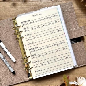 May include: A beige planner with gold-colored rings and a "Debt Tracker" insert. The insert has columns for debt creditor, due date, payments, and new balances. Two markers, a black Sharpie and a white Shuttle Art wet erase marker, are visible.