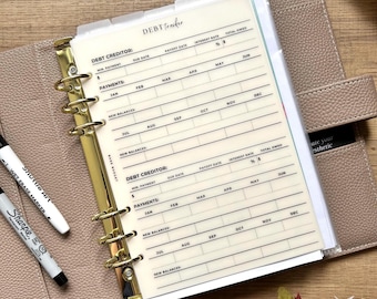 May include: A beige planner with gold-colored rings and a "Debt Tracker" insert. The insert has columns for debt creditor, due date, payments, and new balances. Two markers, a black Sharpie and a white Shuttle Art wet erase marker, are visible.