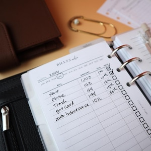 May include: A close-up of a planner open to a "Bills Tracker" page. The page lists bills such as rent, phone, and auto insurance with amounts and due dates. The word "APR" is circled. A brown leather wallet and a gold paper clip are in the background.
