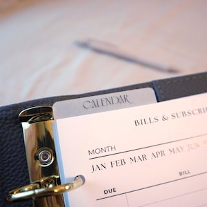 May include: Close-up of an open planner with a gray cover and gold metal rings. The calendar tab is visible, with the word "CALENDAR" printed on it. The planner pages show the months of the year and space for bills and subscriptions.