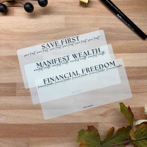 May include: Three translucent cards with the phrases "SAVE FIRST", "MANIFEST WEALTH", and "FINANCIAL FREEDOM" printed in black. A black pen, a wooden die with the number 1, and decorative berries are also visible.