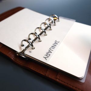 Angled close up of a small silver ring binder in brown croc leather with a matte laminated vellum divider tab labeled JANUARY in thin sans serif letters rounded corners over a white planner page