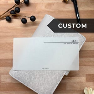 May include: A white leather wallet with a card that reads "RENT ONE MONTH AHEAD" and "BARE BUDGET". The word "CUSTOM" is displayed on a black banner. A black pen and decorative branch are also visible.