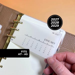 May include: A translucent plastic calendar insert for a ring binder, featuring the month of January 2027 with black text. The insert has gold-toned metal rings and is labeled to fit A7 to A5 sizes. A circular black graphic displays the years 2027, 2028, and 2029.