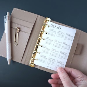 May include: A beige planner with gold accents, open to reveal a 2026 calendar page. A clear pen and gold paperclip rest on the left. The calendar page is held by a hand.