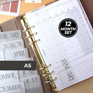 May include: A5 planner with gold-colored rings, open to a monthly calendar page. Includes monthly dividers and a sticker that says "12 MONTH SET". The planner is open to the January page.
