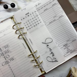 May include: A planner insert with a 2026 calendar for January, featuring a gold binder ring. The insert includes sections for savings and sinking funds, with the word "January" in elegant script. The planner is open, revealing lined pages.