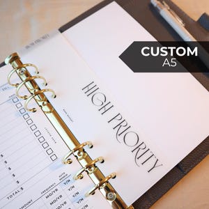May include: Open A5 planner with gold-colored rings and a "HIGH PRIORITY" page. The planner has a black cover and a pen resting on the right side. The text "CUSTOM A5" is on a black arrow-shaped label.