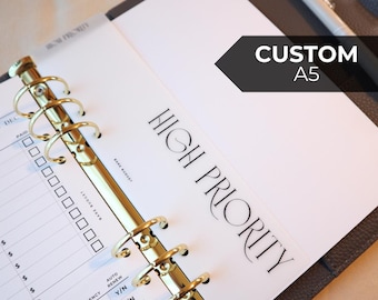 May include: Open A5 planner with gold-colored rings and a "HIGH PRIORITY" page. The planner has a black cover and a pen resting on the right side. The text "CUSTOM A5" is on a black arrow-shaped label.