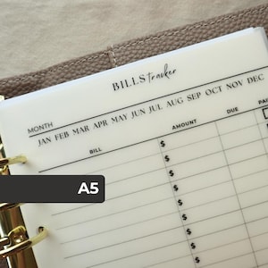 May include: A translucent A5 bills tracker insert with black printed text and lines. The header reads BILLS tracker, followed by months from JAN to DEC. Columns include BILL, AMOUNT with dollar signs, DUE, and PAID, held by gold rings.
