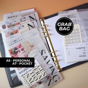 May include: A black six-ring binder containing various translucent planner inserts, including a 2026 calendar, a savings tracker, and a card labeled Generational Wealth. A circular sticker reads Grab Bag, and a card reads Budgeting is my therapy.