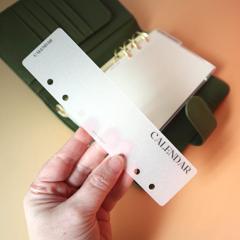 Hand holding a translucent matte vellum bookmark divider tab with punched ring holes and CALENDAR printed in a bold serif font with a green leather ring binder and peach background behind it