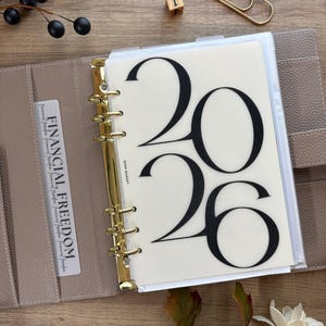 May include: A beige leather planner with gold hardware, open to a page with the large black numbers "2026". A card in the planner reads "FINANCIAL FREEDOM". A gold paperclip and a wooden block with the number "1" are also visible.