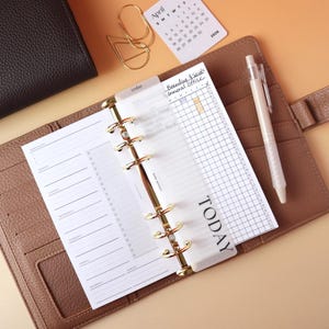 Matte laminated vellum TODAY divider tab in a bold serif font in an open caramel leather ring binder with gold rings next to a weekly planner insert and habit tracker on a peach background with an April 2026 mini calendar card gold paper clips and a pen