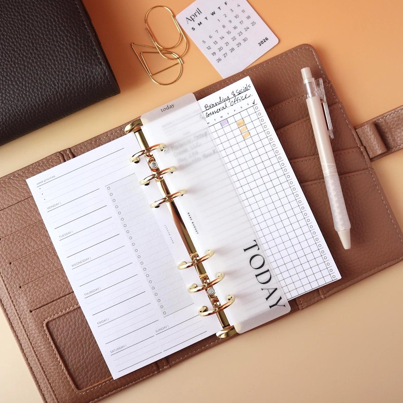 Matte laminated vellum TODAY divider tab in a bold serif font in an open caramel leather ring binder with gold rings next to a weekly planner insert and habit tracker on a peach background with an April 2026 mini calendar card gold paper clips and a pen