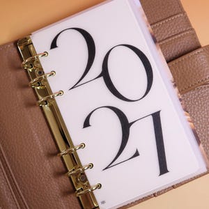 May include: A planner insert made of translucent white material featuring the black printed text 2021. The insert is held by a gold-colored metal six-ring binder mechanism attached to a textured brown leather-like cover with visible stitching.