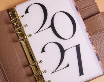 May include: A planner insert made of translucent white material featuring the black printed text 2021. The insert is held by a gold-colored metal six-ring binder mechanism attached to a textured brown leather-like cover with visible stitching.