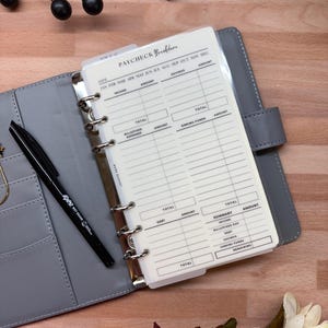 May include: A gray planner with a "Paycheck Breakdown" insert, open to a blank page with sections for income, savings, and expenses. A black pen rests on the planner, and a small wooden die is visible.