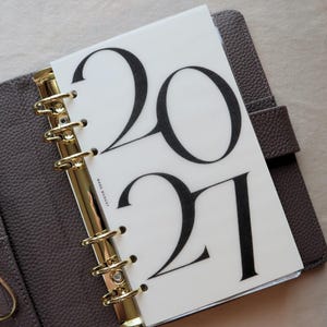 May include: A brown leather-textured binder with a gold-toned six-ring metal mechanism holding a translucent white plastic divider. The divider features the large black printed text 2021 and the smaller vertical text RARE BUDGET.