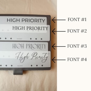 May include: A close-up of a dark gray planner with four different "High Priority" labels in various fonts. The labels are translucent and have a dotted line for the planner's rings. The background is a light beige color.