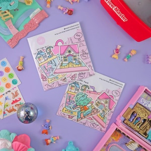 May include: Two sticker packs with pink house illustrations, surrounded by miniature toys and stickers, on a purple surface. The packs feature a pink house with blue doors and windows. Other toys include miniature people and a View-Master.