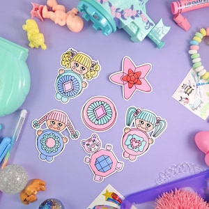 Könnte beinhalten: Eine Sammlung bunter Aufkleber und Spielzeuge auf einer lila Oberfläche. Die Aufkleber zeigen Cartoon-Mädchen, einen Stern und eine Katze, alle in Pastellfarben. Weitere Spielzeuge sind ein rosa Flugzeug, eine Babypuppe und ein Armband.