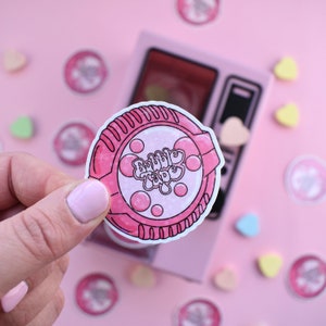 Może przedstawiać: Różowo-biała naklejka z tekstem "Bubble Tape" w retro czcionce. Naklejka ma kształt rolki taśmy i ma różowo-biały wzór z bąbelkami.