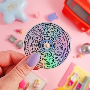 Puede incluir: Un disco compacto holográfico con dibujos y texto en tinta negra. El texto incluye títulos de canciones como "Waterfalls", "Iris", "Mysterious Girl", "Ghetto Superstar", "Never Ever", "Macarena", "Kiss Me", "Swear It Again", "Truly Madly Deeply", "Movin", "I Believe", "What A Girl Wants" y "For My Bestie".