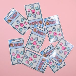 May include: A set of 10 colorful gummy bear stickers with a holographic finish. The stickers are arranged in a grid pattern on a pink background. The stickers feature the text "INDYLION" and "NOTAGMI".