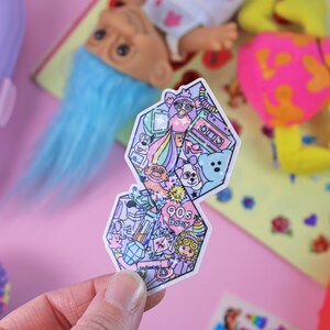 Pu&ograve; includere: Un adesivo a tema baby anni '90. L'adesivo ha la forma di un diamante e presenta una variet&agrave; di icone degli anni '90, tra cui un troll, un Tamagotchi, un nastro per cassette e un arcobaleno.