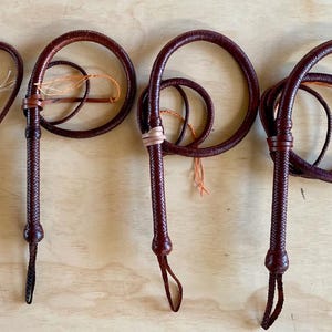 May include: Four brown leather whips with braided handles and looped ends are displayed against a light wood background. The whips vary slightly in design, with some having decorative knots and different loop configurations. The leather appears to be a rich, dark brown colour.
