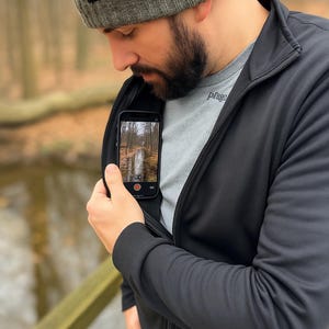 Puede incluir: Un hombre lleva una chaqueta negra con cremallera, una boina gris y una camiseta gris con la palabra "prag" impresa. Está sosteniendo un teléfono inteligente negro en su mano izquierda, y la pantalla muestra una foto de un sendero en el bosque.