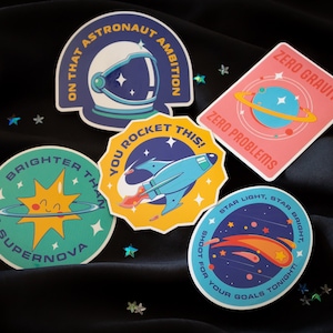 Puede incluir: Cinco pegatinas redondas de colores con ilustraciones y texto sobre el tema del espacio. Las pegatinas representan un cohete, un planeta, una estrella, un sol y un astronauta. El texto en las pegatinas incluye "On that astronaut ambition", "Zero gravity, zero problems", "You rocket this!", "Brighter than a supernova", y "Star light, star bright, shoot for your goals tonight!"