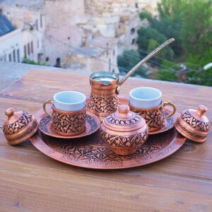 May include: A copper serving tray with intricate floral designs, holding a copper coffee pot, two small copper cups, a sugar bowl, and two lids. The tray is on a wooden table with a blurry background of a town.