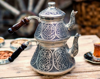 May include: A silver tea set with intricate engraved patterns, featuring a stacked teapot with a lid and two spouts. The teapot has dark wooden handles. The set sits on a weathered wooden table, with a blurred background of a wooden wheel.
