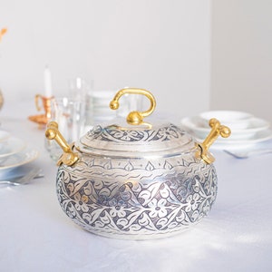 May include: A silver and gold serving dish with an intricate floral design. The dish has two handles and a lid.