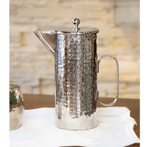 May include: A silver hammered metal pitcher with a lid and a handle. The pitcher has a textured surface and a rounded shape.