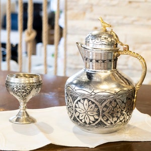 May include: A silver pitcher and goblet set with intricate floral and geometric patterns. The pitcher has a gold-colored handle and a bird-shaped finial on the lid. The set is displayed on a white, scalloped-edge cloth.