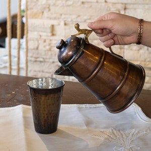 Puede incluir: Una jarra de cobre con un asa de latón se está vertiendo en una taza de cobre sobre un mantel de lino blanco. La jarra y la taza están sobre una mesa de madera.