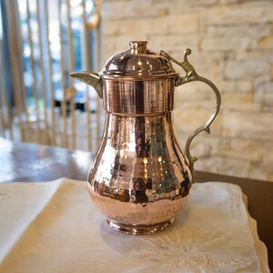 May include: A copper pitcher with a hammered texture, featuring a decorative handle and a lid. The pitcher is set on a white tablecloth with an embroidered design. The background includes a brick wall and a window.