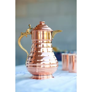 May include: A copper pitcher with a hammered finish and a brass handle and spout. The pitcher has a rounded body and a flared lip.