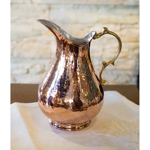 May include: A hammered copper pitcher with a brass handle. The pitcher has a rounded body and a wide mouth.